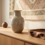 Close-up of a hand-thrown speckled ceramic vase on a carved wooden sideboard with a woven wall hanging in the softly blurred background.